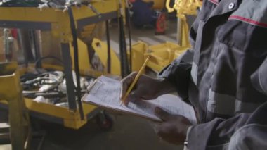 Siyah üretim mühendisinin yan görüntüsü ve beyaz kasketi üretim fabrikasındaki ekipmanların çalışmasını kontrol ederken günlük kontrol listesine not alıyor.