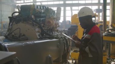 Beyaz kasklı ve üniformalı Afrikalı teknisyenlerin, ağır makine üretim tesisinde büyük ısı motorlarını inceleyip notlar aldıkları orta ölçekli bir fotoğraf.