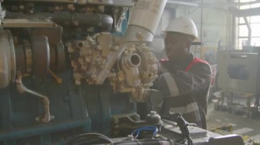 Afrika kökenli Amerikalı erkek fabrika işçisinin üretim fabrikasındaki büyük içten yanmalı motorun kasnaklarını tamir veya kontrol ederken orta ölçekli görüntüsü.