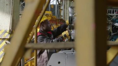 Afrika kökenli Amerikalı erkek fabrika işçisinin kasketli ve üniformalı kamera görüntüleri. Makine fabrikasındaki traktörün iç patlama motoru üzerinde çalışan makaralar.