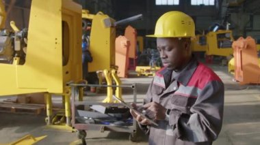 Tablet kullanan ve motor montajı yapan ağır makine üretim tesisinde motor çerçevelerinin yanında duran Afrikalı Amerikalı erkek yöneticinin orta derece yavaşlaması
