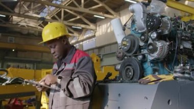 Sarı kasklı ve tulumlu Afrikalı Amerikalı erkek teknisyenin, büyük patlama motorunun makaralarını teftiş ederken ve sonra da makine fabrikasındaki tablete bilgi yazarken orta boy görüntüsü.
