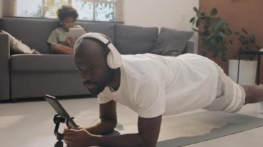 Oğlu dijital tablette videolar izlerken, babasının yoga dersini takip ederken, kulaklık takıp, tahta pozisyonda oturduğu tam bir fotoğraf.