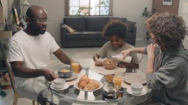 Üç kişilik bir ailenin oturma odasında otururken birlikte kahvaltı ettiği orta boy bir fotoğraf.