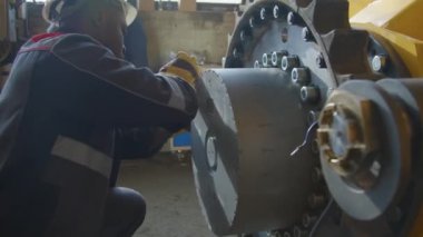 Afrikalı Amerikalı fabrika işçisinin, ağır makine üretim tesisinde ingiliz anahtarı kullanarak traktör iskeletinde fındık ezerken yan çekimi.