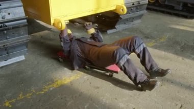 Yüksek açılı, sarı kasketli ve üniformalı siyah erkek işçinin tırtıl traktör iskeletinin altında yuvarlanırken ve makine fabrikasında vida ya da fındık vidalarken görüntüsü.