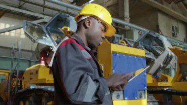 Afrikalı Amerikalı fabrika teknisyeninin sarı başlık takıp tablete veri yazarken ağır makine fabrikasındaki ekipman üretimini analiz ederken orta kavisli görüntüsü.