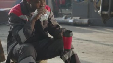 Erkek Afro-Amerikan fabrika işçisinin öğle yemeği molasında makine fabrikasında tek başına yemek ve içkinin tadını çıkarırken orta derece çekimleri.