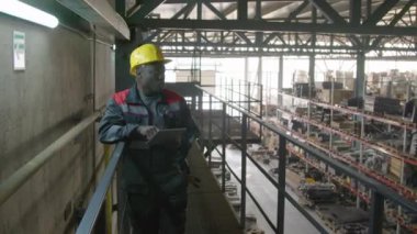 Orta boy Afro-Amerikan erkek depo müdürü sarı kasklı ve üniformalı yukarıdan mal stoklarını kontrol ediyor.