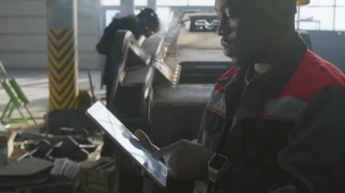 Siyah erkek üretim mühendisinin, sarı baret ve tulumlu, makine fabrikasındaki kaynak alanında tablet üzerinde çalışırken yan yatış görüntüsü.