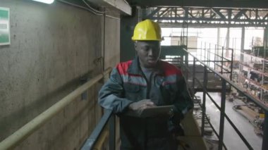 Siyah şapkalı ve tulumlu Afro-Amerikan erkek depo müdürünün tablete bilgi yazarken yukarıdan depolama ögelerini kontrol ederken orta boy görüntüsü.