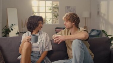 Genç evli bir çiftin kanepede oturup, sıcak içecekler içip birbirleriyle konuştuğu orta boy bir fotoğraf.