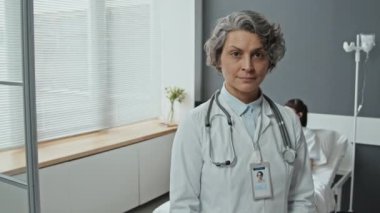 Beyaz önlükle hastane koğuşunda dikilip hasta arka planda yatarken kameraya bakan beyaz kadın doktorun seçmeli odak portresi.