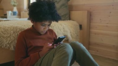 Orta boy Afro-Amerikan çocuk portresi gündelik giysiler giymiş yerde oturmuş akıllı telefondan internette sörf yapıyor.