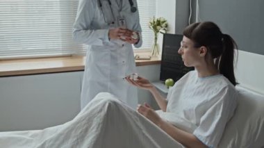 Doktorun ilaç getirdiği ve kadın hastasının hastane yatağında otururken ilaç aldığı orta boy bir fotoğraf.