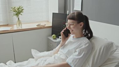 Hastane önlüklü, yatakta telefonla konuşan beyaz bir kadının orta boy fotoğrafı.