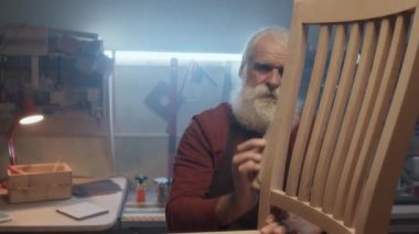 Orta yaşlı marangoz, gri saçlı, gömlekli ve önlüklü ahşap sandalyeli.