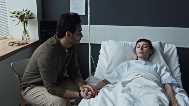 ベッドで意識不明の彼のコーカサスガールフレンドの手を握っている病院区に座っている中東男性のミドルショット — ストック動画