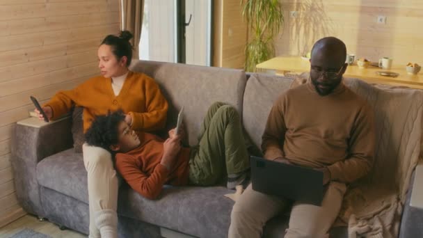 现代多元种族的父母和孩子们坐在沙发上 用智能手机 数字平板电脑和笔记本电脑放松身心 — 图库视频影像