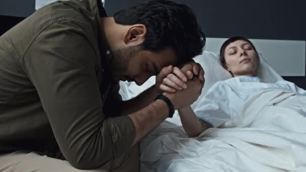 昏睡状態で病院のベッドに横たわる彼の最愛の手を保持する心配中東の男性の選択的な焦点中クローズアップショット — ストック動画