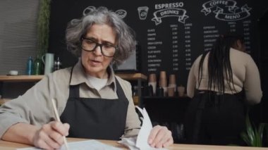 Genç meslektaşı arka planda çalışırken, yaşlı küçük işletme sahibinin hesapları uzlaştırırken orta ölçekli bir fotoğrafı.