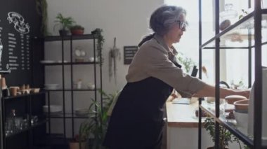 Genç meslektaşı tezgahın arkasında müşterileri beklerken yaşlı kadın kahve dükkanı çalışanı bisküvilerle kavanozları yerleştiriyor.