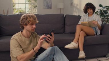 Genç evli bir çift, iyi ışıklandırılmış oturma odasında birlikte otururken ayrı aktiviteler yapıyorlar. Cep telefonuyla mesajlaşıyorlar ve kanepede kitap okuyorlar.