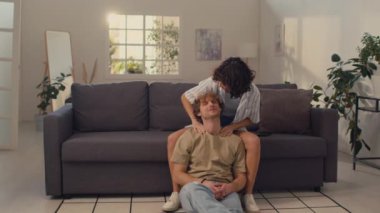 Siyah kıvırcık saçlı genç bir kadın kanepede oturup erkek arkadaşına omuz masajı yapıyor.
