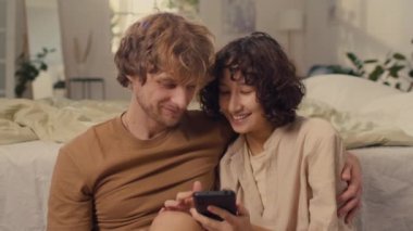 Genç mutlu çiftin evlerinde oturmuş telefondaki resimlere bakarken orta boy fotoğrafı.