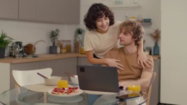 Genç beyaz adam mutfakta oturmuş laptopuyla çalışırken kahvaltı yapıyor ve kız arkadaşı da ona arkadan sarılıyor.