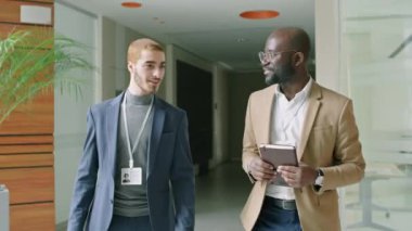 Resmi takım elbiseler altında isim kartı takan iki erkek iş yeri koridorunda yürüyorlar sohbet ediyorlar.