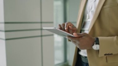 Resmi takım elbiseli siyahi bir adamın dijital tabletini kullanarak koridorda yürüyüp mesajlaşırken orta ölçekli yakın çekimi.