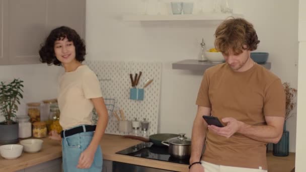 彼女のボーイフレンドが彼の携帯電話をスクロールしながらキッチンで暗いカーリーヘアクリーニングで若い女性のメディアショット — ストック動画
