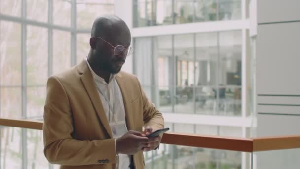 ベージュスーツのアフリカ系アメリカ人のビジネスマンは 彼のスマートフォンを使用してカーテンウォールの前に屋内で立っている名前バッジを身に着けています — ストック動画