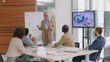 Kadın teknoloji şirketinin CEO 'su beyaz tahtadaki finansal çizelgelere işaret ediyor ve ofiste yapılan iş toplantısında çok ırklı çalışanlara sunum yapıyor.