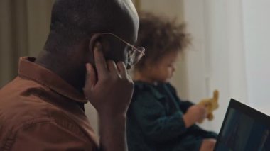 Siyah adamın kablosuz kulaklık taktığı seçici odak noktası kızı arka planda otururken iş arkadaşlarıyla online görüşme yapması.