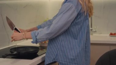 Açık mavi tişörtlü, tanınmayan sarışın kadın mutfakta durmuş çırpılmış yumurta yapıyor.