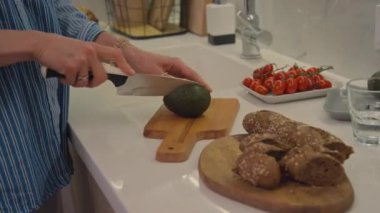 Beyaz, sarışın kadın mutfakta duruyor. Avokado, domates ve ekmekle yemek hazırlıyor. Bıçak ve kesim tahtası kullanıyor.