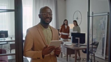 Şeftali rengi resmi takım elbise giyen, elinde dijital tablet olan, kadın meslektaşlarının önünde dikilip kameraya bakan Afro-Amerikan bir adamın portresi.