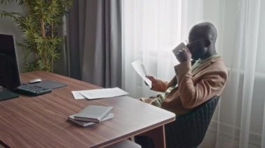 Resmi takım elbiseli genç CEO iş yerinde oturmuş sıcak kahve içerken belgelere bakıyor.
