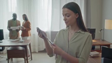 Genç beyaz kadın ressamın portresi toplantı odasının ortasında duruyor, elinde farklı renklerde bir şablon tutuyor, kameraya bakıyor.