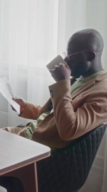 Resmi takım elbiseli genç CEO iş yerinde oturmuş sıcak kahve içerken belgelere bakıyor.