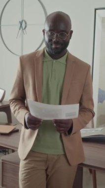 Afro-Amerikan CEO 'su ofisinde durmuş yeşil gömlekli, resmi bej bir kostüm giyiyor, elinde belgelerle kameraya bakıyor ve gülümsüyor.
