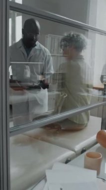Yeni kol protezini test eden kıvırcık saçlı genç bir kadının camından dikey çekim. Doktor telefonda konuşurken kadın hemşirenin yardımıyla.