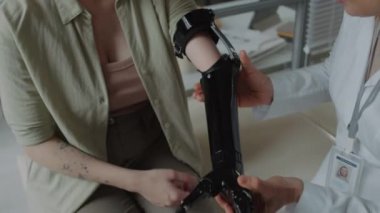 Ampütasyonlu genç bayana yardım eden tıbbi personel muayene koltuğunda oturup plastik kol protezi takıyor.