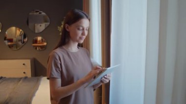 Stüdyonun penceresinin yanında duran bej tişörtlü genç bir kadının portresi dijital tablete, sonra da kameraya bakıyor.