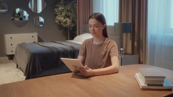 暗い柔らかい色で装飾された彼女の寝室に座っている若い白人学生 デジタルタブレットを使用して勉強 — ストック動画