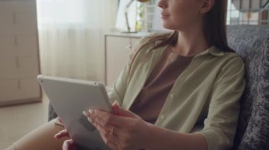 Genç beyaz kadın oturma odasında koltukta rahatça oturuyor, pencereye bakıyor, sonra e-kitap okumaya başlıyor.