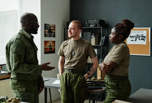 Three young intercultural students in military uniform having discussion of main points of training while standing in front of camera