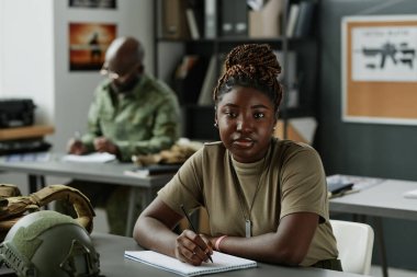 Sıradan giyinen Afrikalı Amerikalı bir kız öğrenci askeri ekipmanlarla masada oturuyor ve ders notları alırken kameraya bakıyor.